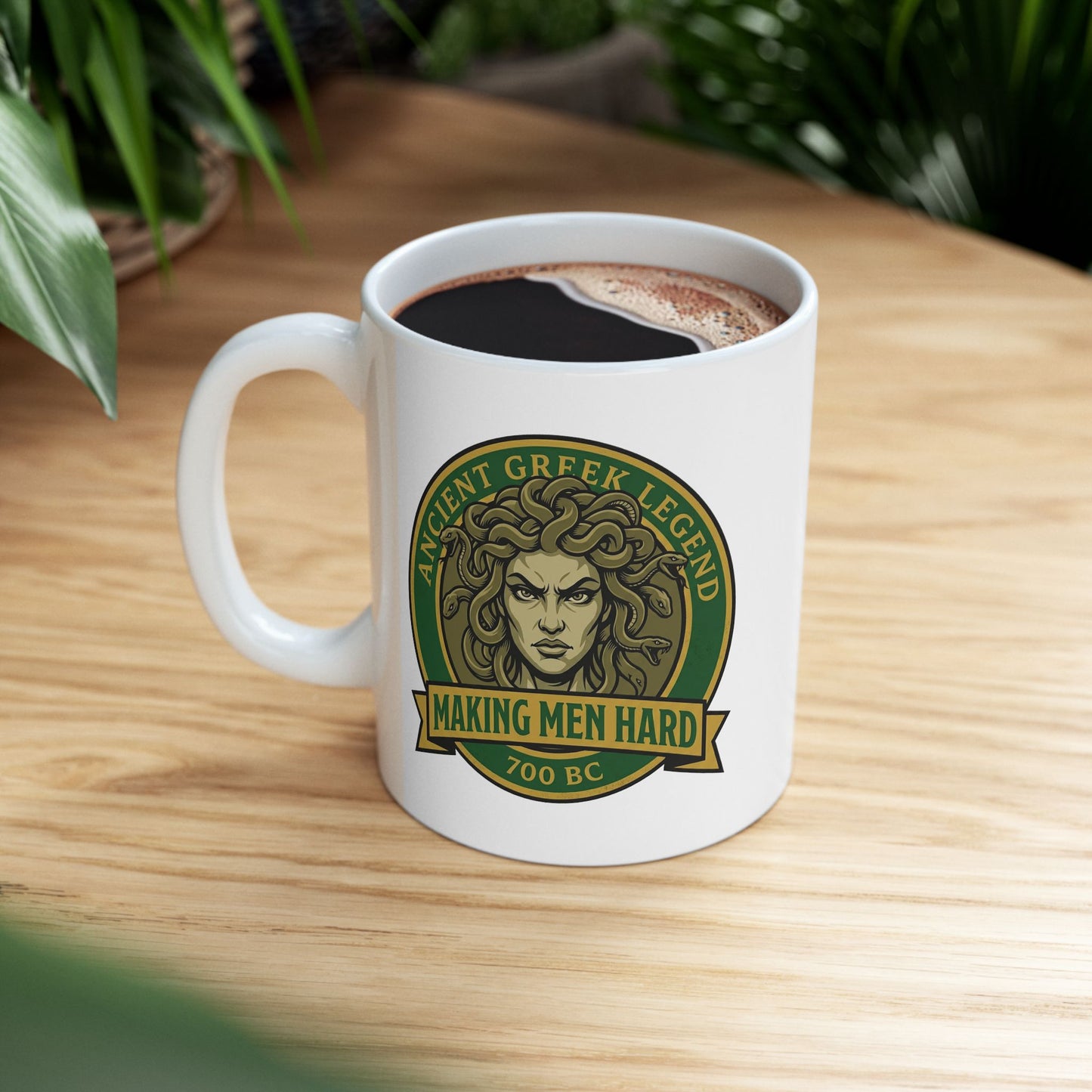 Funny Greek mythology mug design featuring a bold vintage badge with Medusa's stern face surrounded by writhing snakes. The circular emblem reads "Ancient Greek Legend" across the top and "Making Men Hard" on a banner below, with "700 BC" at the bottom. The design uses classic green, gold, and olive tones in a retro emblem style, combining mythology with humorous wordplay.