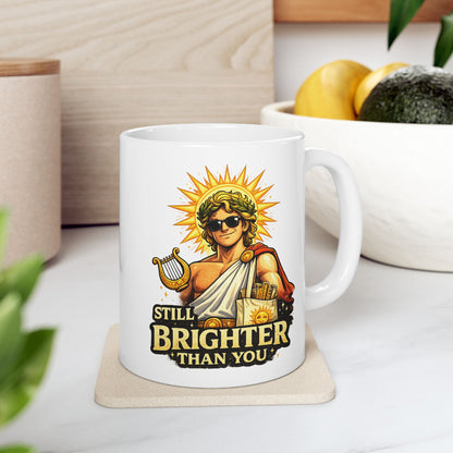 White ceramic mug with a humorous, vibrant illustration of the Greek god Apollo wearing dark sunglasses and a laurel wreath, with a glowing golden sunburst behind his head. He holds a golden lyre and wears a toga with a red cape. Below the smirking god, the cheeky text reads "STILL BRIGHTER THAN YOU" in a bold, weathered yellow font.