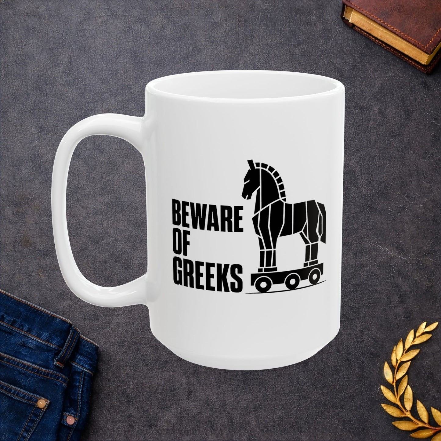 White ceramic mug with a minimalist black-and-white graphic featuring a stylized, geometric silhouette of the Trojan Horse on a wheeled platform. To the left of the horse, the text "BEWARE OF GREEKS" is written vertically in a bold, stacked, black sans-serif font. The design uses clean lines and high contrast on a plain background, offering a modern, minimalist take on the classic Greek mythological tale.