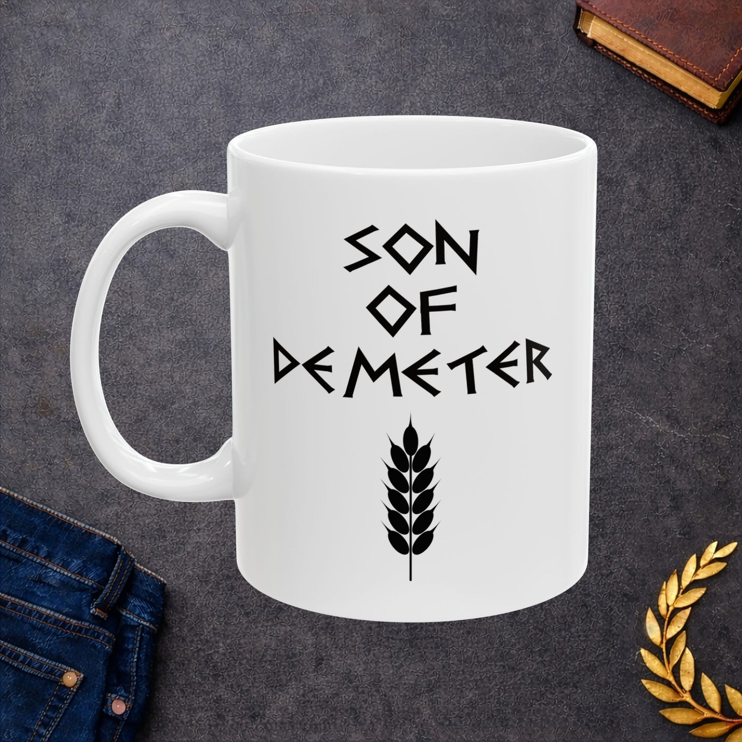 White ceramic mug featuring a minimalist, Greek-inspired graphic in black ink on a white background. The design reads 'SON OF DEMETER' in a stylized, angular font, positioned above a clean, line-art graphic of a bundle of harvested wheat.
