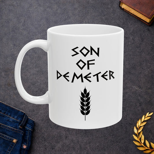 White ceramic mug featuring a minimalist, Greek-inspired graphic in black ink on a white background. The design reads 'SON OF DEMETER' in a stylized, angular font, positioned above a clean, line-art graphic of a bundle of harvested wheat.