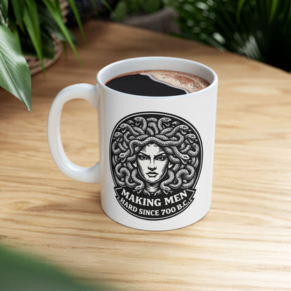 White ceramic mug with a bold black-and-white circular emblem featuring the head of Medusa with a mass of detailed, writhing snakes for hair. Below the illustration, a curved banner contains the humorous double-entendre "MAKING MEN HARD SINCE 700 B.C." written in a heavy, white serif font. The design uses a high-contrast, woodcut-inspired aesthetic with intricate line work against a dark background.
