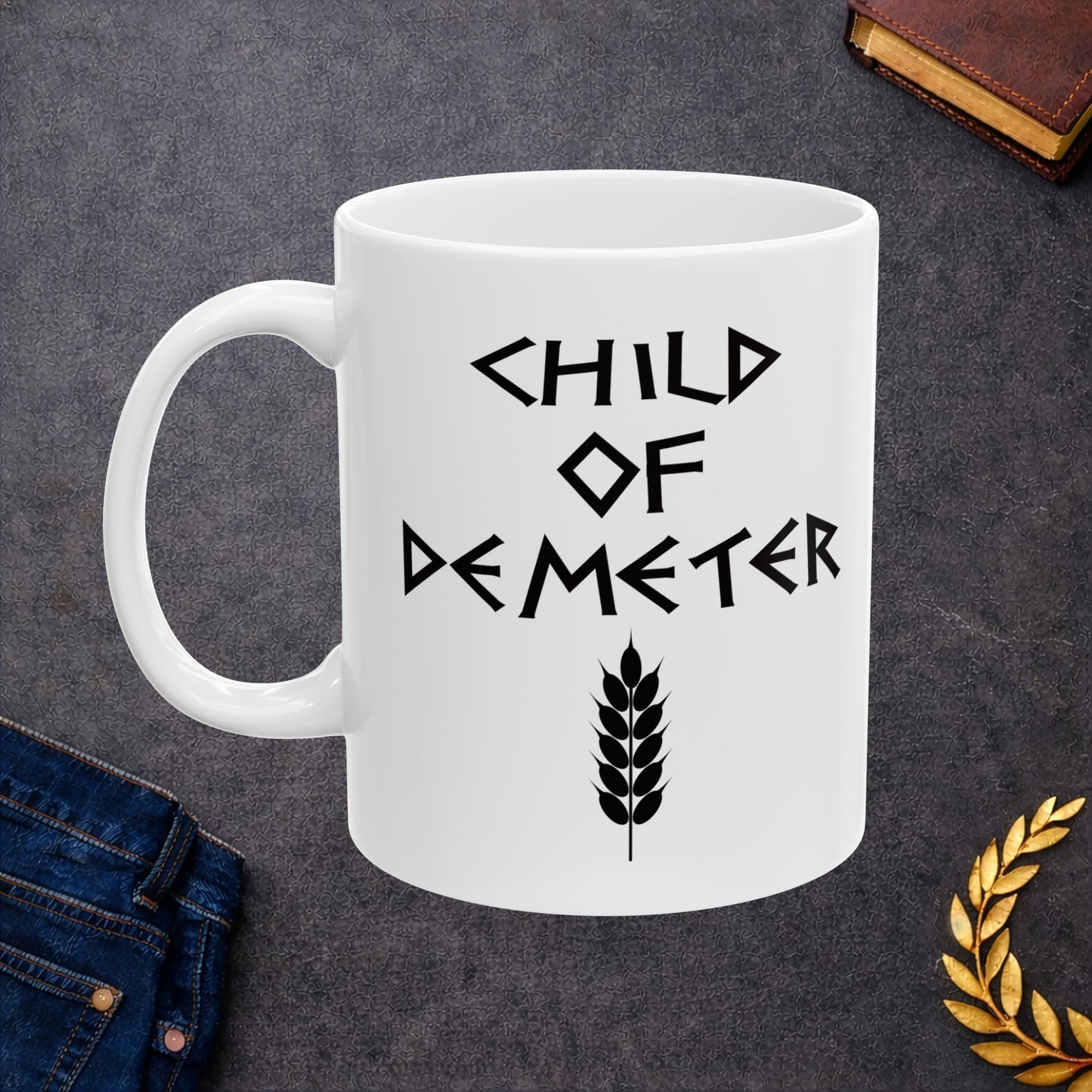 White ceramic mug featuring a minimalist, Greek-inspired graphic in black ink on a white background. The design reads 'CHILD OF DEMETER' in a stylized, angular font, positioned above a clean, line-art graphic of a bundle of harvested wheat.