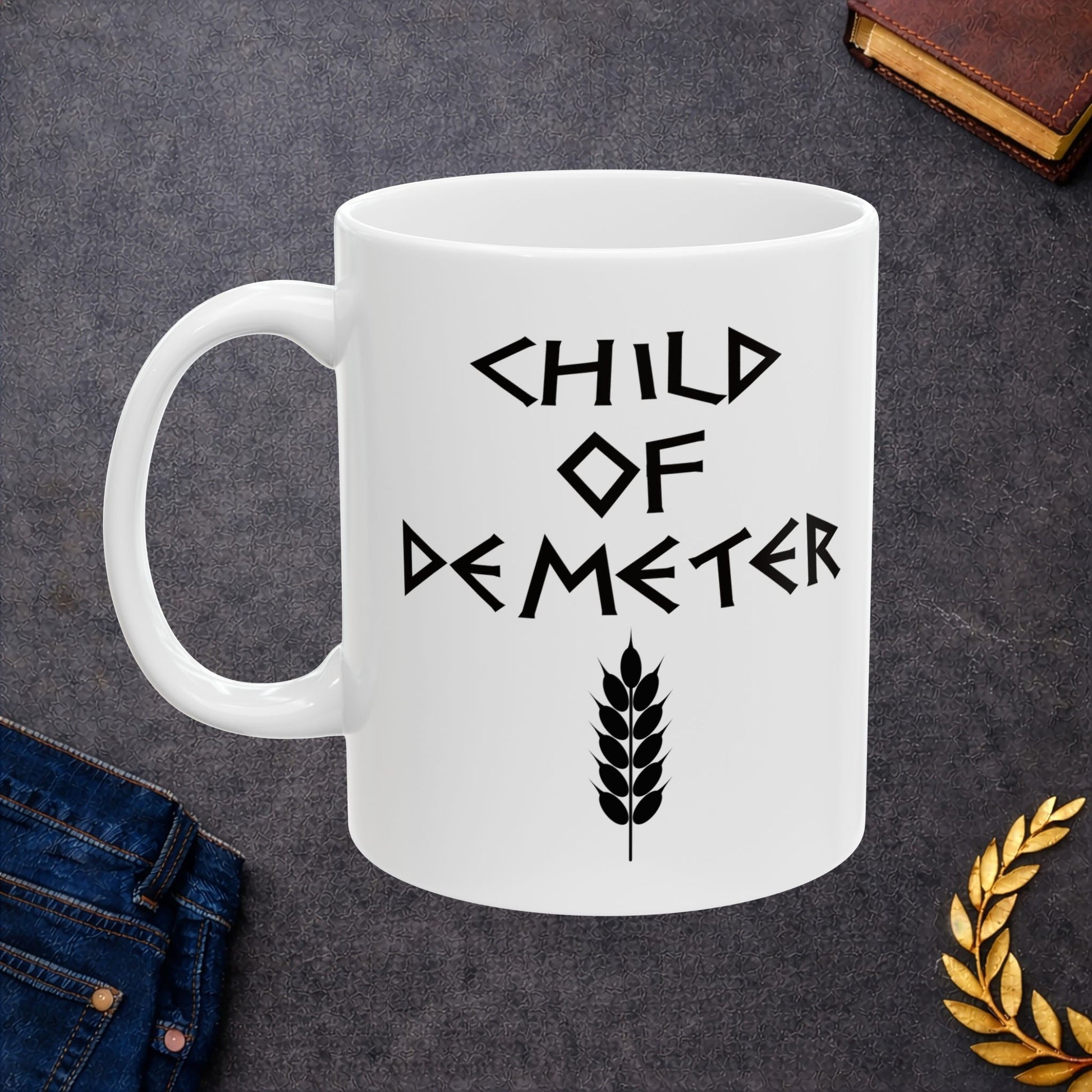 White ceramic mug featuring a minimalist, Greek-inspired graphic in black ink on a white background. The design reads 'CHILD OF DEMETER' in a stylized, angular font, positioned above a clean, line-art graphic of a bundle of harvested wheat.