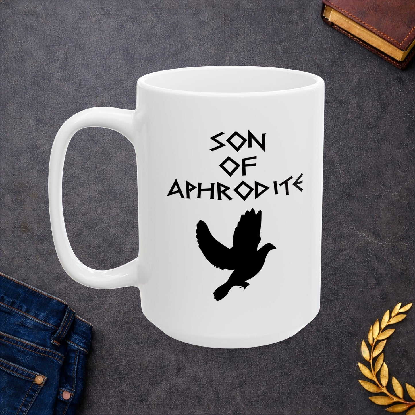 White ceramic mug featuring a minimalist, Greek-inspired graphic in black ink on a white background. The design reads 'SON OF APHRODITE' in a stylized, angular font, positioned above a clean, line-art graphic of a dove in flight.