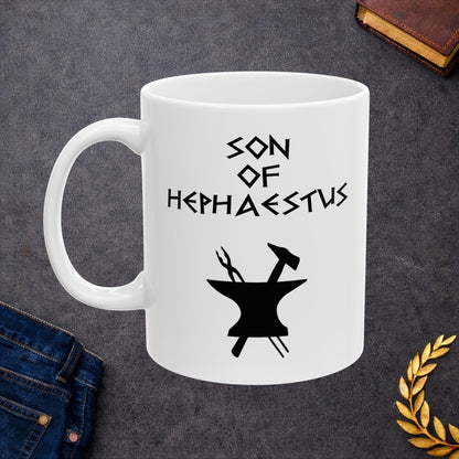 A minimalist black graphic for a white ceramic mug. The design features a central silhouette of a blacksmith's anvil with a hammer and tongs crossed behind it. Above the tools, the text "SON OF HEPHAESTUS" is written in a sharp, angular font inspired by ancient Greek inscriptions. The entire illustration is rendered in a clean, solid black style.