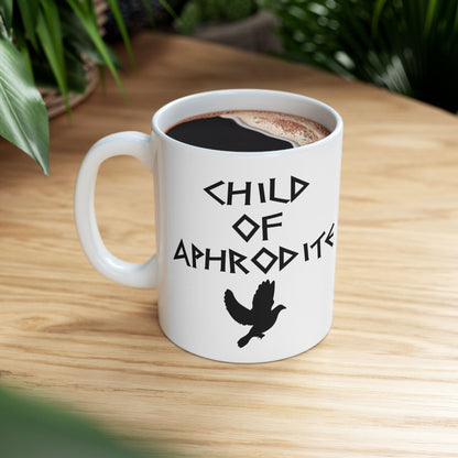 White ceramic mug featuring a minimalist, Greek-inspired graphic in black ink on a white background. The design reads 'CHILD OF APHRODITE' in a stylized, angular font, positioned above a clean, line-art graphic of a dove in flight.