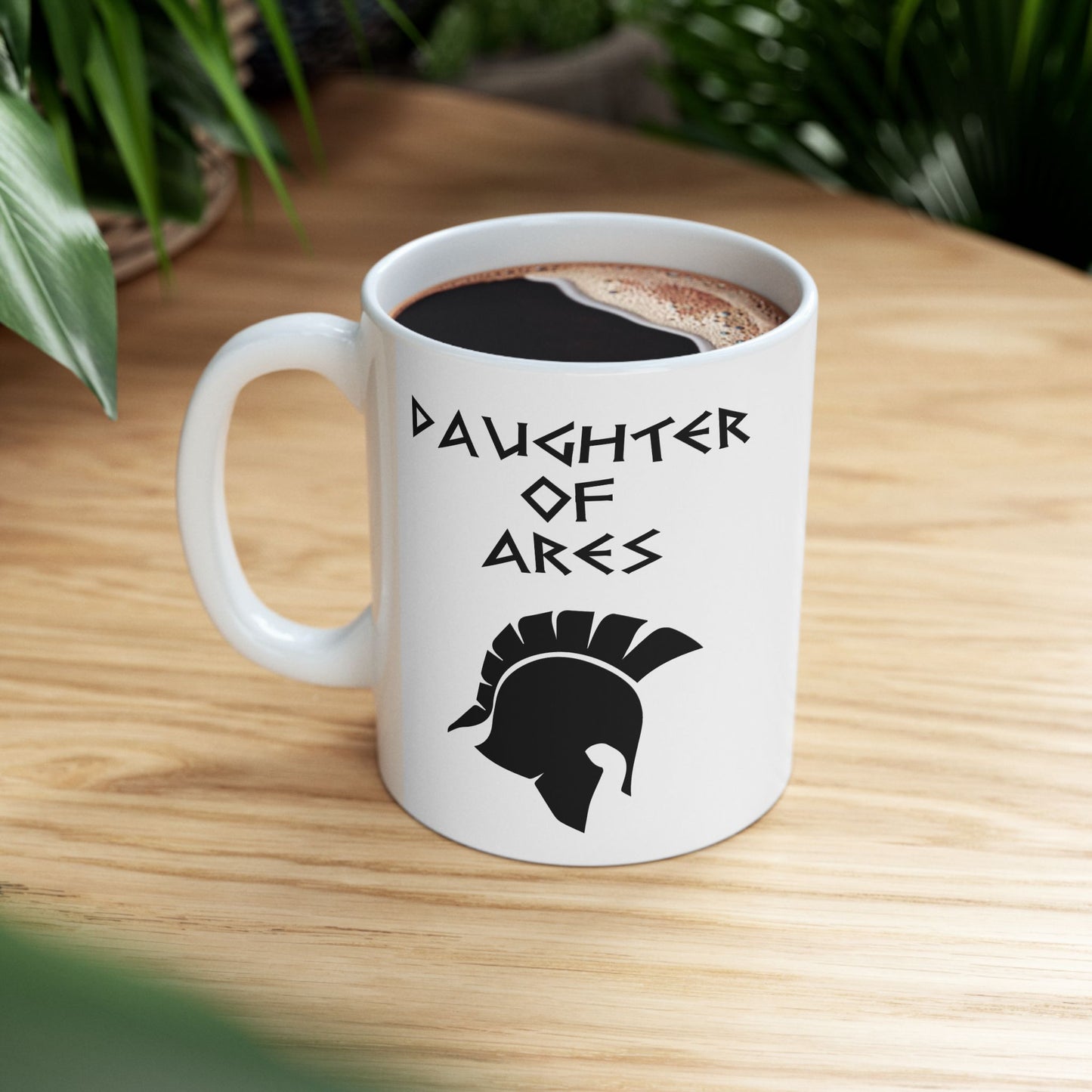White ceramic mug featuring a minimalist, Greek-inspired graphic in black ink on a white background. The design reads 'DAUGHTER OF ARES' in a stylized, angular font, positioned above a clean, line-art graphic of a Spartan-style plumed warrior helmet.