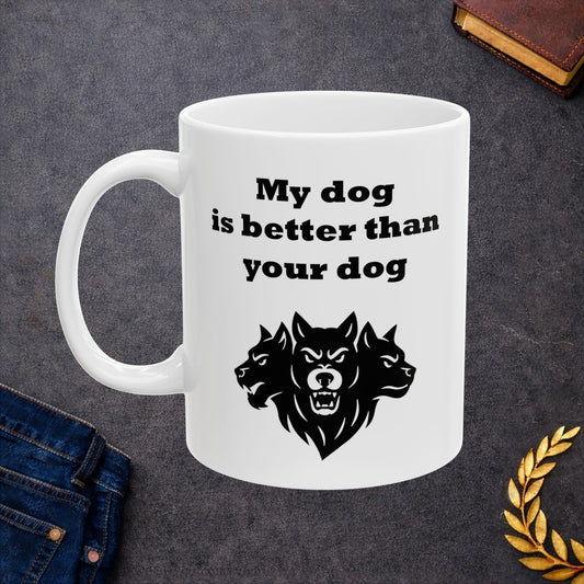 White ceramic mug with a minimalist black-and-white graphic featuring a bold, three-headed Cerberus illustration. The hound of the underworld is shown with snarling expressions and sharp teeth. Above the heads, the phrase "My dog is better than your dog" is written in a heavy, black slab-serif font. The design is a clean, modern take on Greek mythology with a humorous twist.