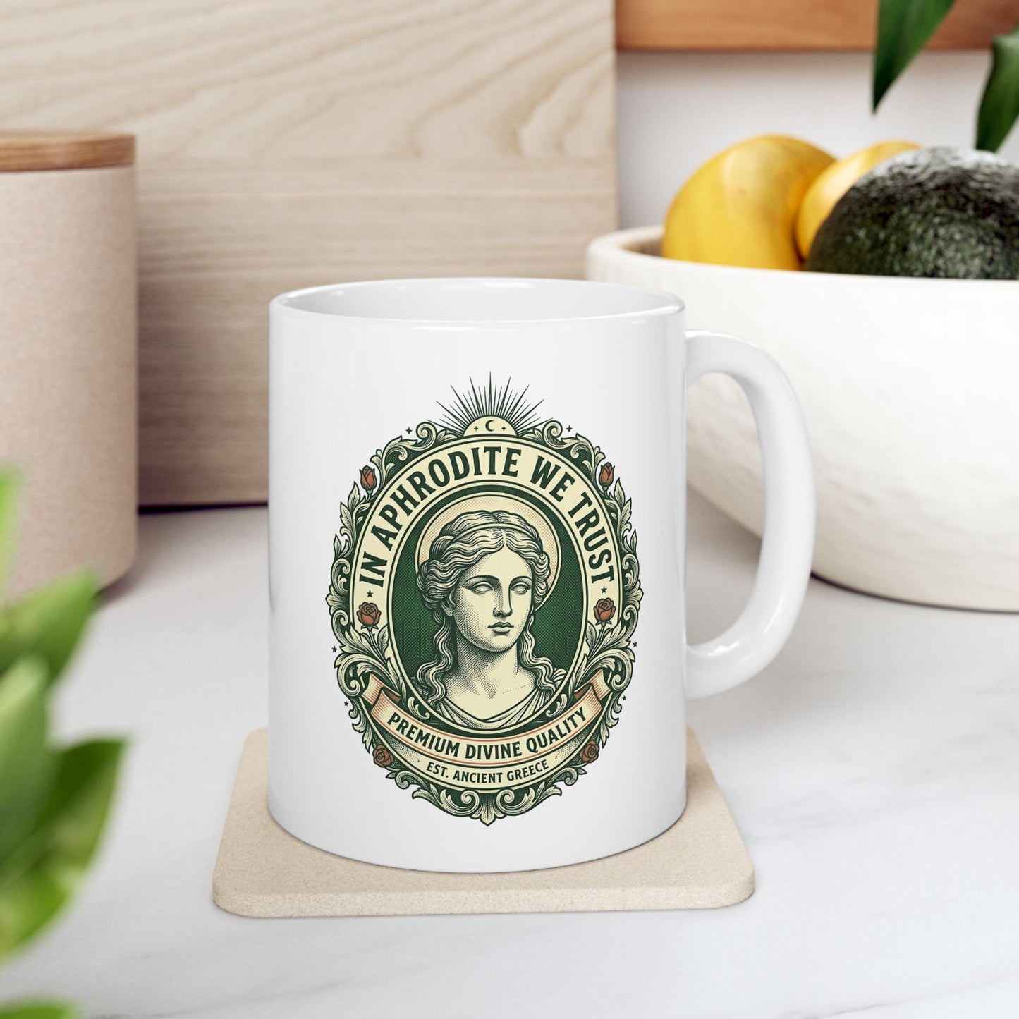 White ceramic mug with a vintage, currency-inspired emblem featuring a detailed bust of the goddess Aphrodite. The design is framed by an ornate oval border with roses and scrolls. The text "IN APHRODITE WE TRUST" arches above, while banners below read "PREMIUM DIVINE QUALITY" and "EST. ANCIENT GREECE." The artwork uses a classic green and cream color palette with a textured, engraved aesthetic.