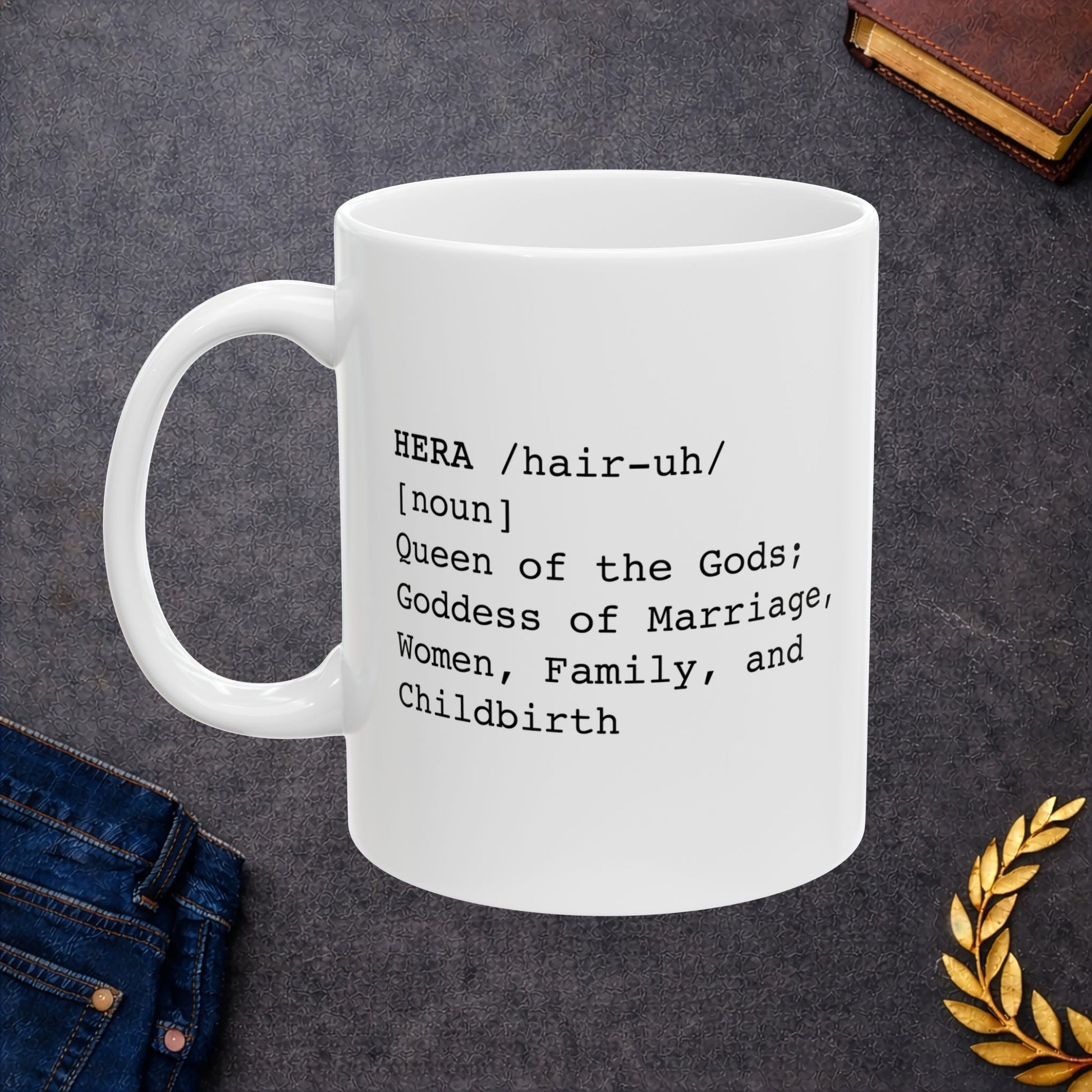 White ceramic mug with a minimalist black text design styled like a dictionary entry. It displays the word "HERA" in capital letters, followed by the pronunciation guide "/hair-uh/" and the part of speech "[noun]". The definition underneath reads: "Queen of the Gods; Goddess of Marriage, Women, Family, and Childbirth." The layout uses a clean, modern typewriter-style font.
