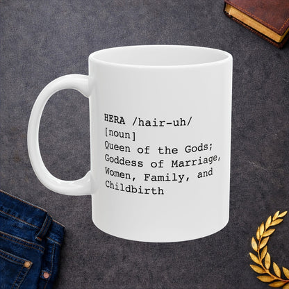 White ceramic mug with a minimalist black text design styled like a dictionary entry. It displays the word "HERA" in capital letters, followed by the pronunciation guide "/hair-uh/" and the part of speech "[noun]". The definition underneath reads: "Queen of the Gods; Goddess of Marriage, Women, Family, and Childbirth." The layout uses a clean, modern typewriter-style font.