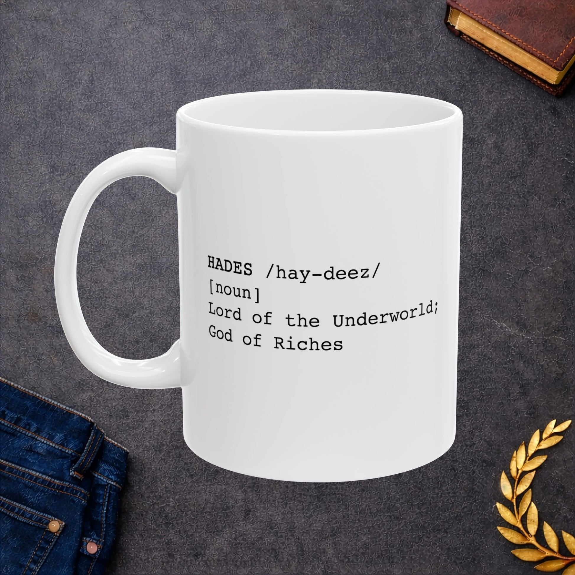 A minimalist graphic for a white ceramic mug featuring black typewriter-style text. The design is formatted like a dictionary entry for "HADES," including the phonetic pronunciation "/hay-deez/," the word class "[noun]," and the definition: "Lord of the Underworld; God of Riches.
