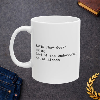 A minimalist graphic for a white ceramic mug featuring black typewriter-style text. The design is formatted like a dictionary entry for "HADES," including the phonetic pronunciation "/hay-deez/," the word class "[noun]," and the definition: "Lord of the Underworld; God of Riches.