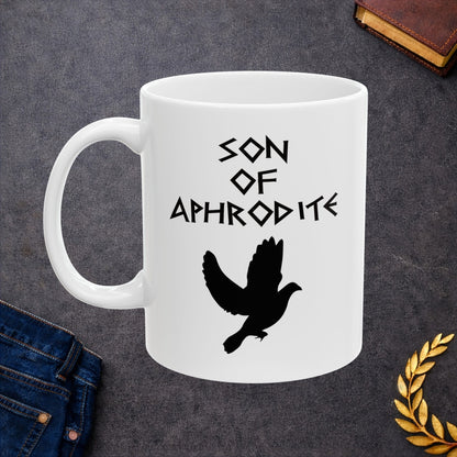 White ceramic mug featuring a minimalist, Greek-inspired graphic in black ink on a white background. The design reads 'SON OF APHRODITE' in a stylized, angular font, positioned above a clean, line-art graphic of a dove in flight.