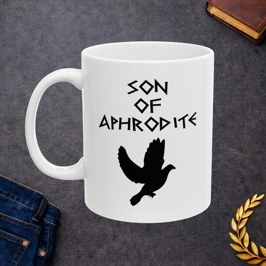 White ceramic mug featuring a minimalist, Greek-inspired graphic in black ink on a white background. The design reads 'SON OF APHRODITE' in a stylized, angular font, positioned above a clean, line-art graphic of a dove in flight.