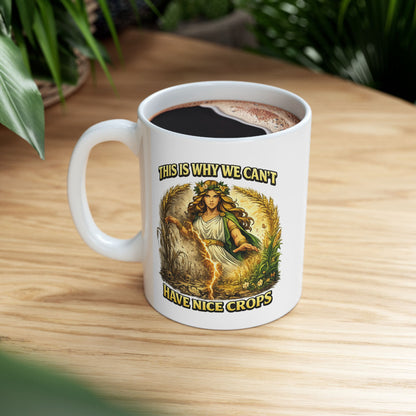 White ceramic mug with a dramatic illustration of Demeter, the Greek goddess of agriculture, standing over a split field. A glowing lightning strike cracks down the center, dividing flourishing, green crops on the right from dead, withered stalks and skulls on the left. Arching text above and below reads "THIS IS WHY WE CAN'T HAVE NICE CROPS" in a bold, weathered yellow font. The design has an intense, mythical comic art style.