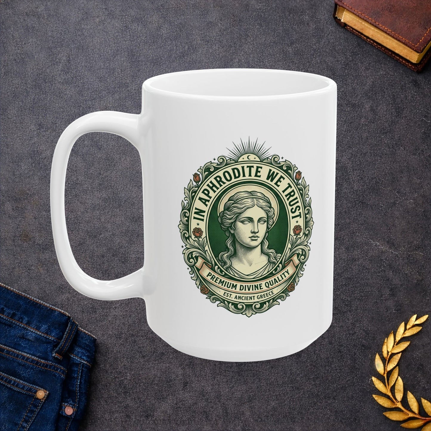 White ceramic mug with a vintage, currency-inspired emblem featuring a detailed bust of the goddess Aphrodite. The design is framed by an ornate oval border with roses and scrolls. The text "IN APHRODITE WE TRUST" arches above, while banners below read "PREMIUM DIVINE QUALITY" and "EST. ANCIENT GREECE." The artwork uses a classic green and cream color palette with a textured, engraved aesthetic.