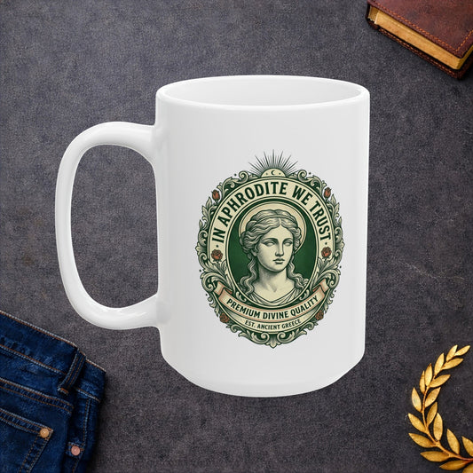 White ceramic mug with a vintage, currency-inspired emblem featuring a detailed bust of the goddess Aphrodite. The design is framed by an ornate oval border with roses and scrolls. The text "IN APHRODITE WE TRUST" arches above, while banners below read "PREMIUM DIVINE QUALITY" and "EST. ANCIENT GREECE." The artwork uses a classic green and cream color palette with a textured, engraved aesthetic.