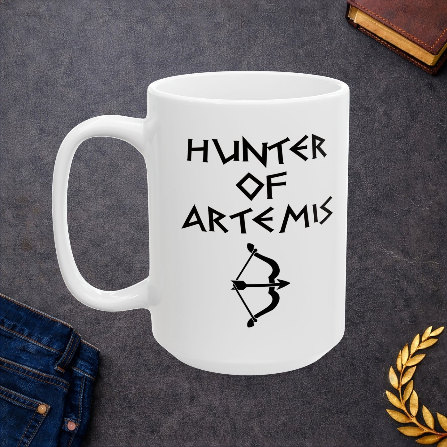 White ceramic mug featuring a minimalist, Greek-inspired graphic in black ink on a white background. The design reads 'HUNTER OF ARTEMIS' in a stylized, angular font, positioned above a clean, line-art graphic of a bow and a notched arrow.