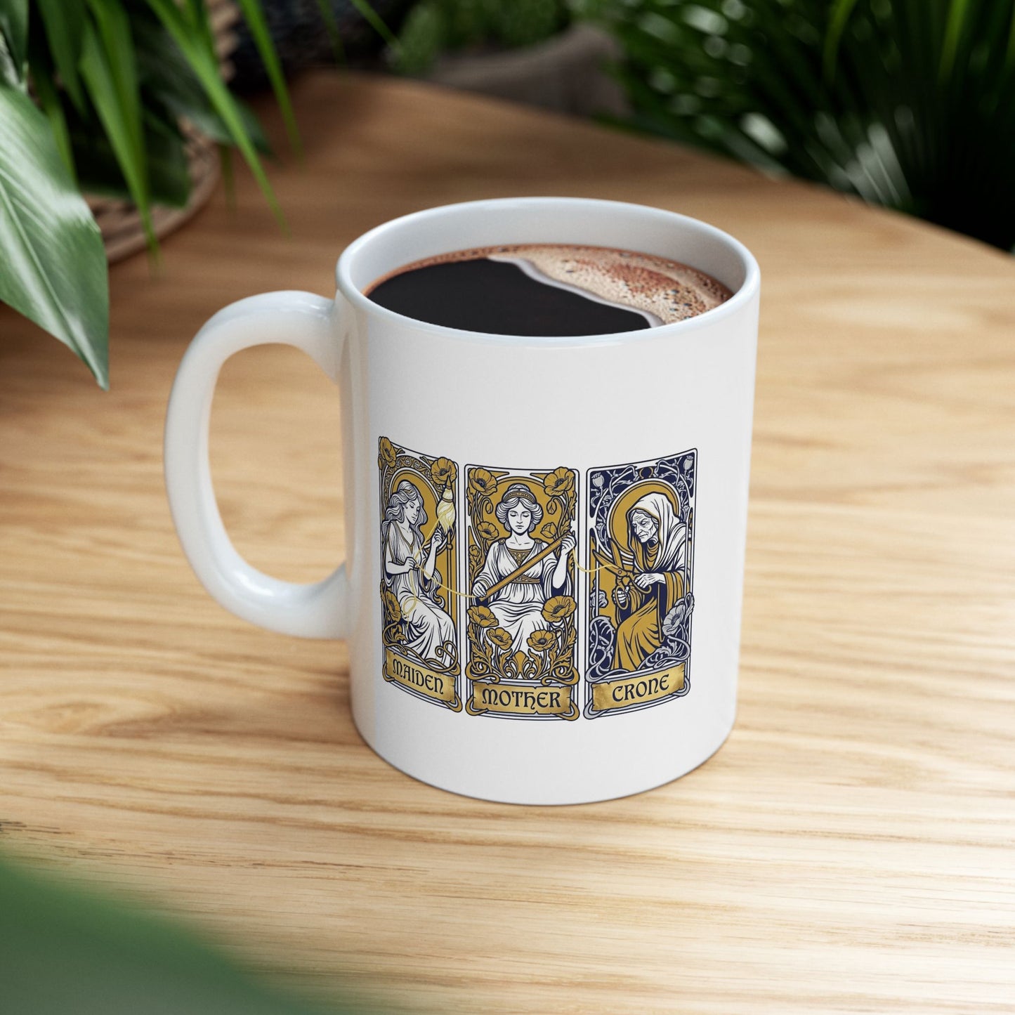 White ceramic mug with Three tarot-style cards depicting the triple goddess archetype: Maiden, Mother, and Crone. The maiden holds a bow surrounded by yellow poppies, the mother wears a crown with a sword, and the crone wears hooded robes with a staff. Each rendered in Art Nouveau style with ornate decorative borders, gold halos, and floral flourishes. Labels appear at card bases. Celebrates the feminine divine in all life stages and cyclical transformation.