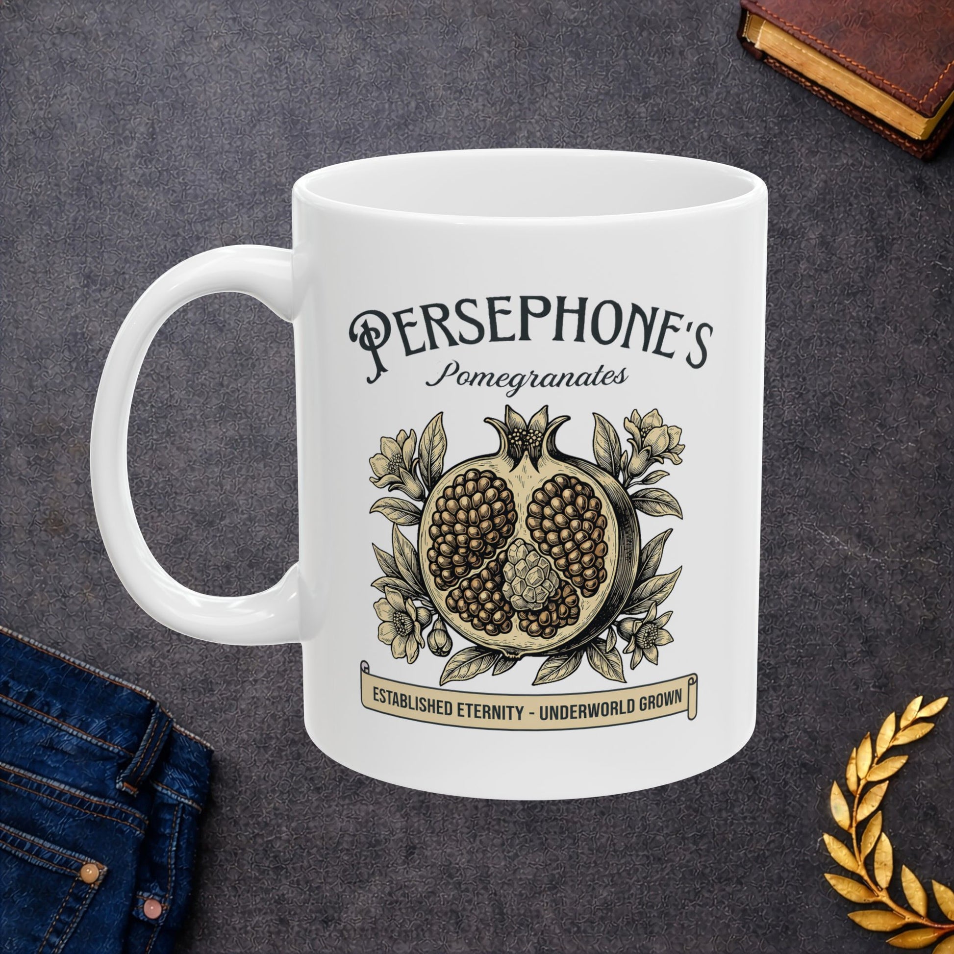 White ceramic mug with a vintage-style botanical illustration of a halved pomegranate showing its seeds, surrounded by delicate pomegranate flowers and leaves. Above the fruit, the text "PERSEPHONE'S Pomegranates" is written in a mix of serif and script fonts. A banner at the bottom reads "ESTABLISHED ETERNITY - UNDERWORLD GROWN." The design features an elegant, woodcut aesthetic in muted earthy tones.