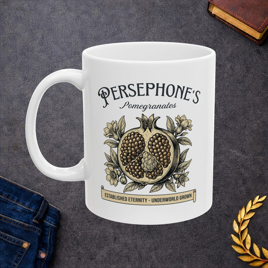 White ceramic mug with a vintage-style botanical illustration of a halved pomegranate showing its seeds, surrounded by delicate pomegranate flowers and leaves. Above the fruit, the text "PERSEPHONE'S Pomegranates" is written in a mix of serif and script fonts. A banner at the bottom reads "ESTABLISHED ETERNITY - UNDERWORLD GROWN." The design features an elegant, woodcut aesthetic in muted earthy tones.
