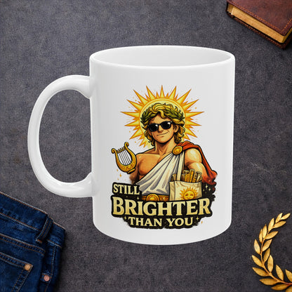 White ceramic mug with a humorous, vibrant illustration of the Greek god Apollo wearing dark sunglasses and a laurel wreath, with a glowing golden sunburst behind his head. He holds a golden lyre and wears a toga with a red cape. Below the smirking god, the cheeky text reads "STILL BRIGHTER THAN YOU" in a bold, weathered yellow font.