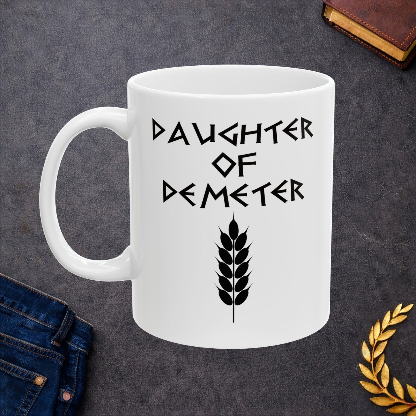 White ceramic mug featuring a minimalist, Greek-inspired graphic in black ink on a white background. The design reads 'DAUGHTER OF DEMETER' in a stylized, angular font, positioned above a clean, line-art graphic of a bundle of harvested wheat.