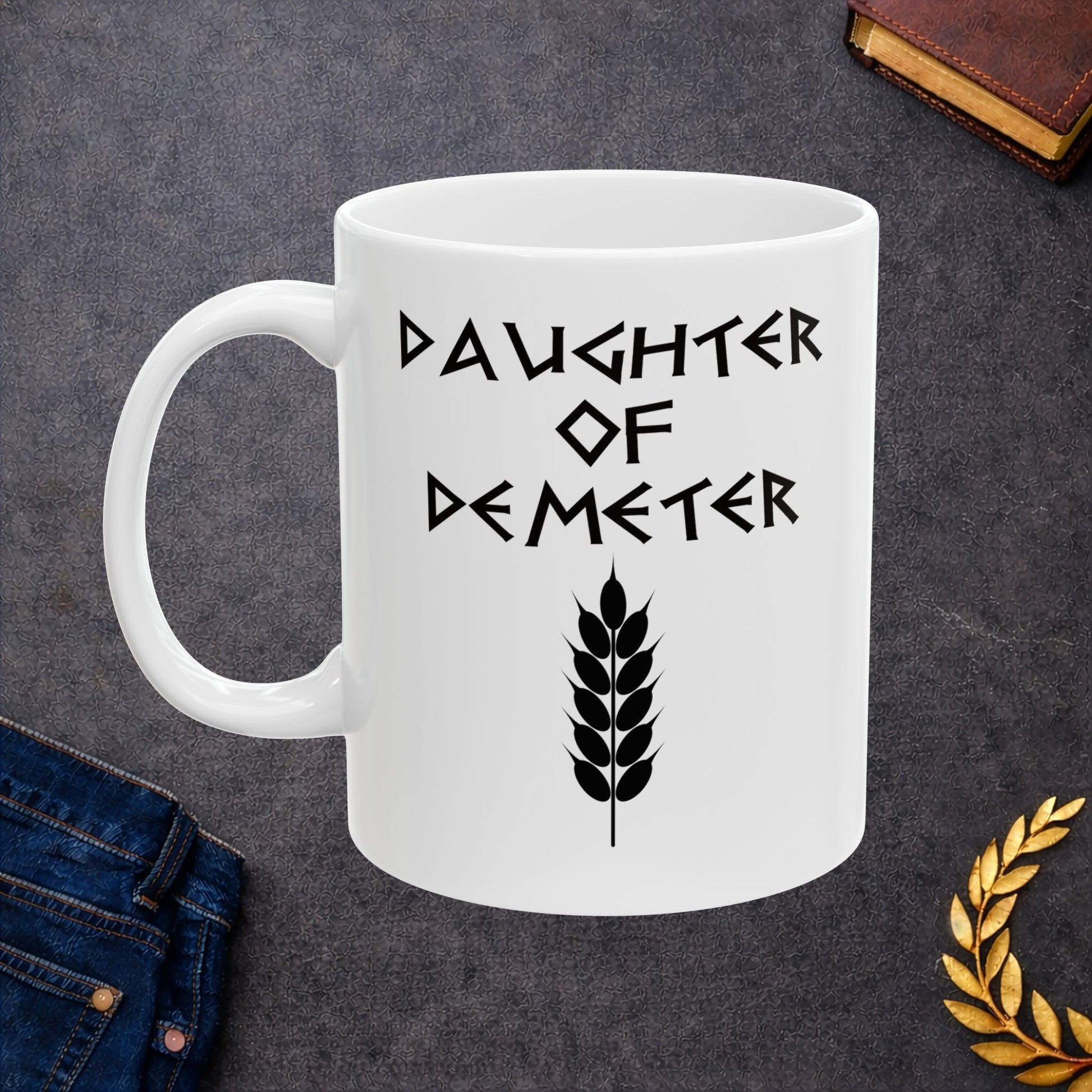 White ceramic mug featuring a minimalist, Greek-inspired graphic in black ink on a white background. The design reads 'DAUGHTER OF DEMETER' in a stylized, angular font, positioned above a clean, line-art graphic of a bundle of harvested wheat.
