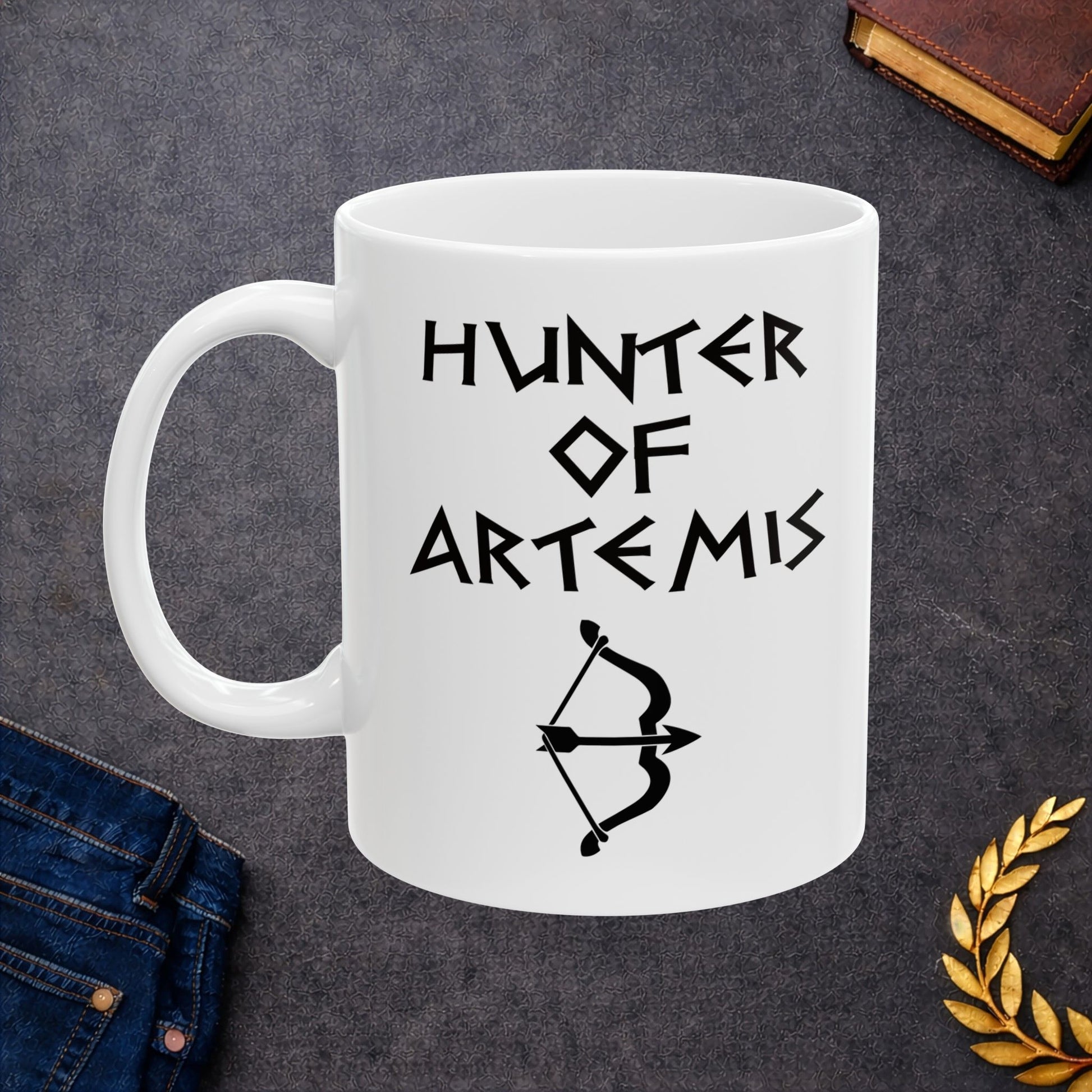 White ceramic mug featuring a minimalist, Greek-inspired graphic in black ink on a white background. The design reads 'HUNTER OF ARTEMIS' in a stylized, angular font, positioned above a clean, line-art graphic of a bow and a notched arrow.