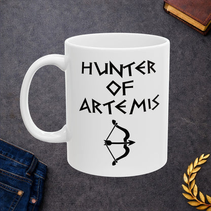 White ceramic mug featuring a minimalist, Greek-inspired graphic in black ink on a white background. The design reads 'HUNTER OF ARTEMIS' in a stylized, angular font, positioned above a clean, line-art graphic of a bow and a notched arrow.