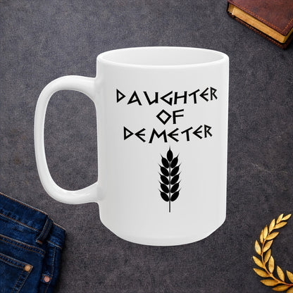 White ceramic mug featuring a minimalist, Greek-inspired graphic in black ink on a white background. The design reads 'DAUGHTER OF DEMETER' in a stylized, angular font, positioned above a clean, line-art graphic of a bundle of harvested wheat.