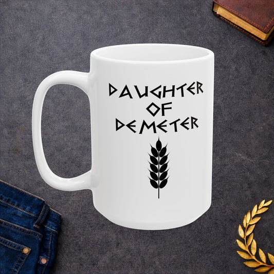 White ceramic mug featuring a minimalist, Greek-inspired graphic in black ink on a white background. The design reads 'DAUGHTER OF DEMETER' in a stylized, angular font, positioned above a clean, line-art graphic of a bundle of harvested wheat.