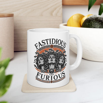 White ceramic mug with a Humorous design combining 'Fast and Furious' with Greek mythology on a white ceramic mug. Text reads 'FASTIDIOUS AND FURIOUS' in distressed lettering surrounding three faces of Medusa in black and white engraving style. Detailed serpents with individual scales form her hair. Greek key pattern borders in orange frame the composition with decorative sun motifs and a small snake head icon at center. Blends classical mythology with contemporary pop culture humor.