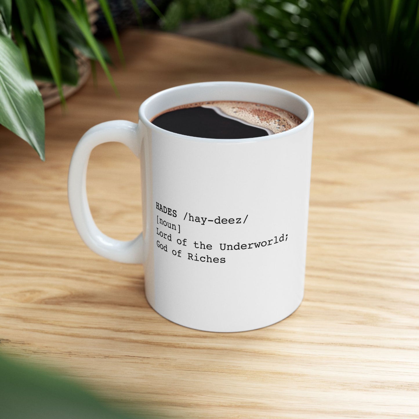 A minimalist graphic for a white ceramic mug featuring black typewriter-style text. The design is formatted like a dictionary entry for "HADES," including the phonetic pronunciation "/hay-deez/," the word class "[noun]," and the definition: "Lord of the Underworld; God of Riches.