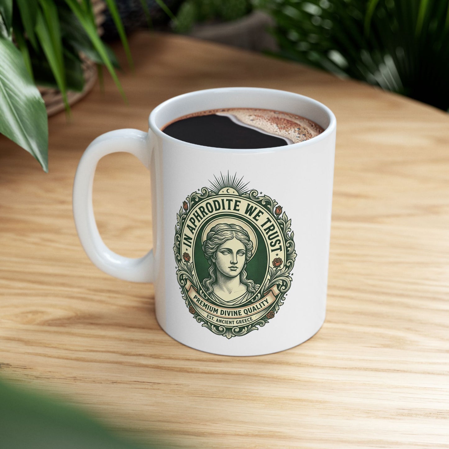 White ceramic mug with a vintage, currency-inspired emblem featuring a detailed bust of the goddess Aphrodite. The design is framed by an ornate oval border with roses and scrolls. The text "IN APHRODITE WE TRUST" arches above, while banners below read "PREMIUM DIVINE QUALITY" and "EST. ANCIENT GREECE." The artwork uses a classic green and cream color palette with a textured, engraved aesthetic.