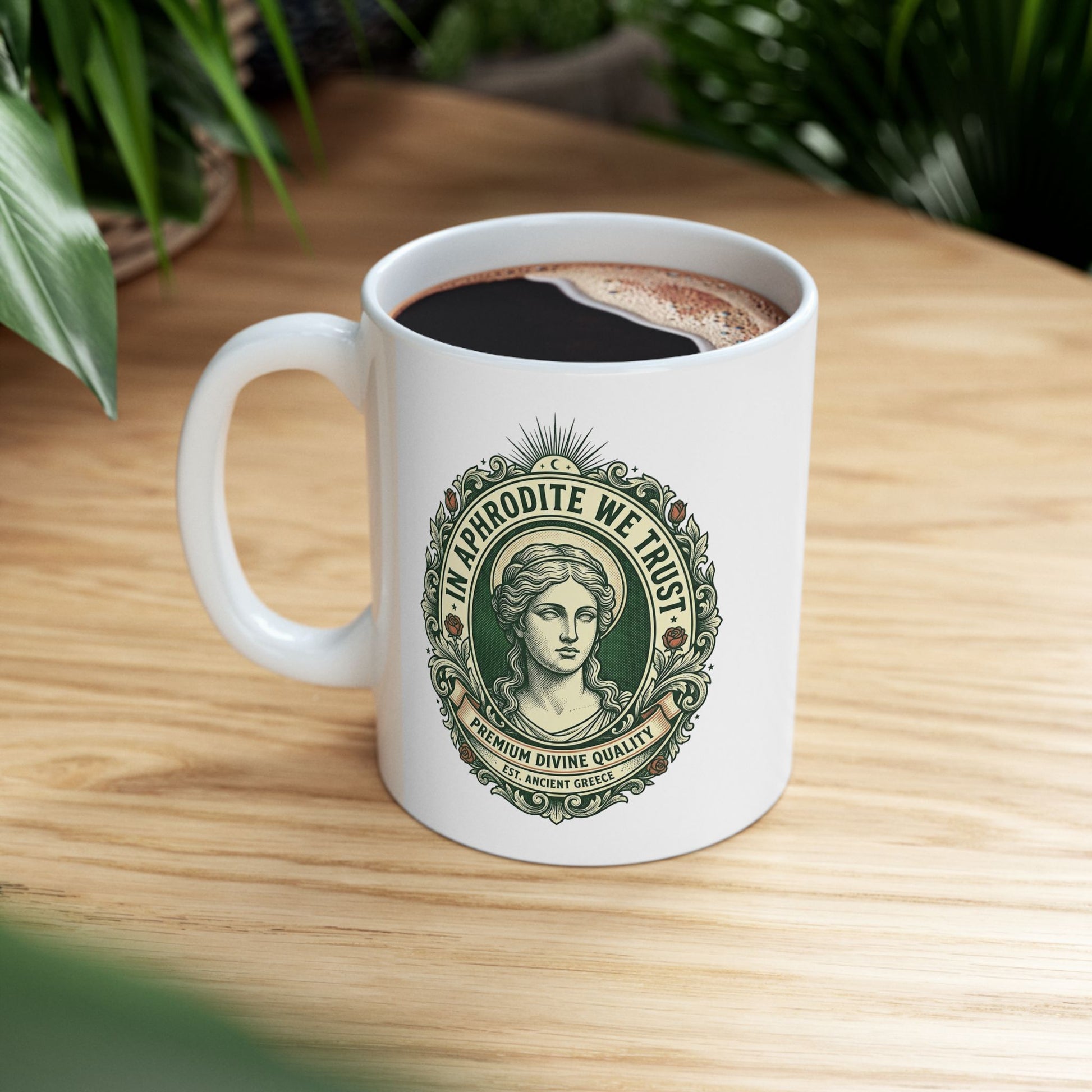 White ceramic mug with a vintage, currency-inspired emblem featuring a detailed bust of the goddess Aphrodite. The design is framed by an ornate oval border with roses and scrolls. The text "IN APHRODITE WE TRUST" arches above, while banners below read "PREMIUM DIVINE QUALITY" and "EST. ANCIENT GREECE." The artwork uses a classic green and cream color palette with a textured, engraved aesthetic.
