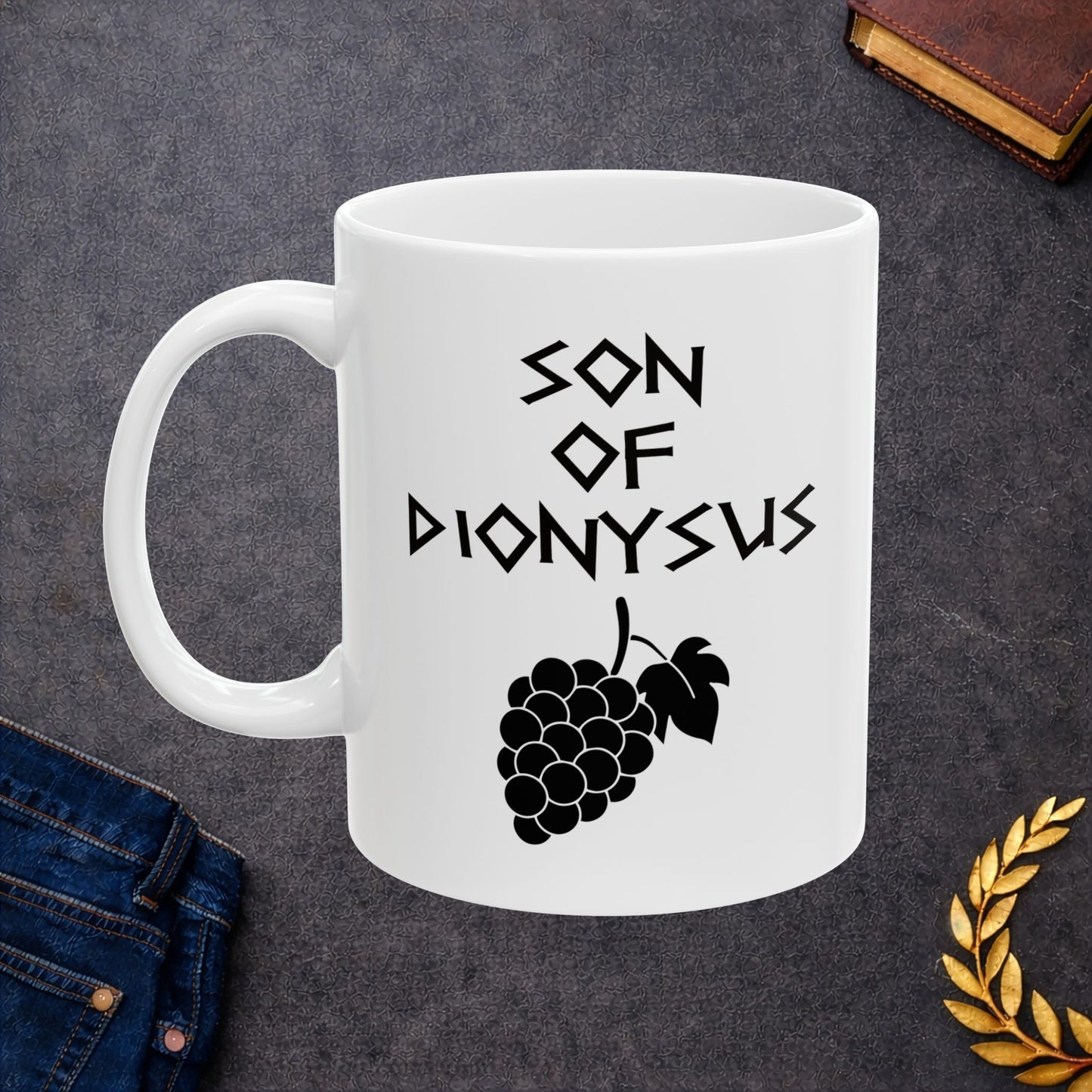 White ceramic mug featuring a minimalist, Greek-inspired graphic in black ink on a white background. The design reads 'SON OF DIONYSUS' in a stylized, angular font, positioned above a clean, line-art graphic of a cluster of grapes hanging from a vine with leaves.