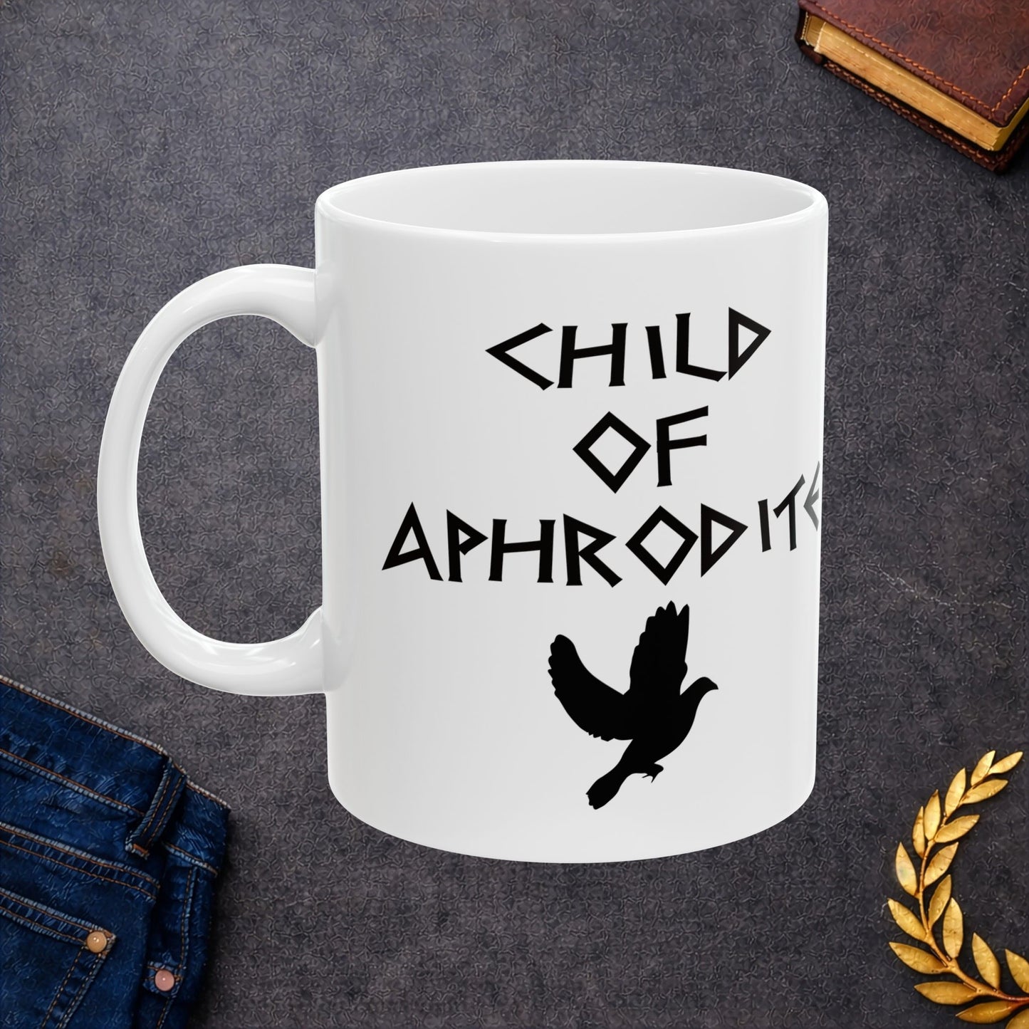 White ceramic mug featuring a minimalist, Greek-inspired graphic in black ink on a white background. The design reads 'CHILD OF APHRODITE' in a stylized, angular font, positioned above a clean, line-art graphic of a dove in flight.