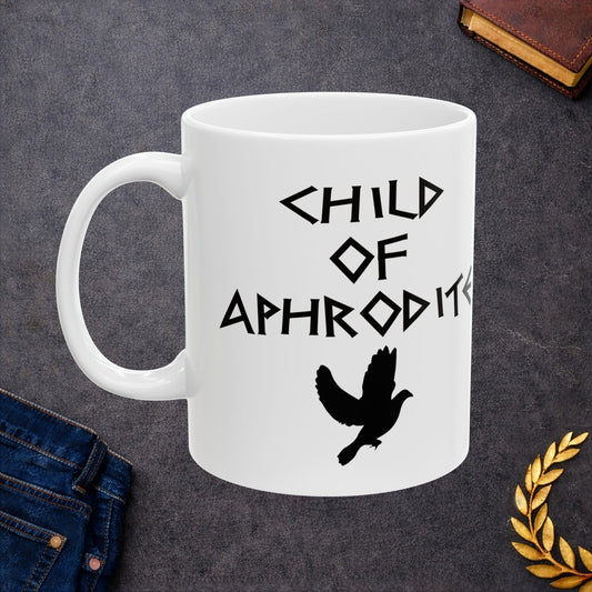 White ceramic mug featuring a minimalist, Greek-inspired graphic in black ink on a white background. The design reads 'CHILD OF APHRODITE' in a stylized, angular font, positioned above a clean, line-art graphic of a dove in flight.
