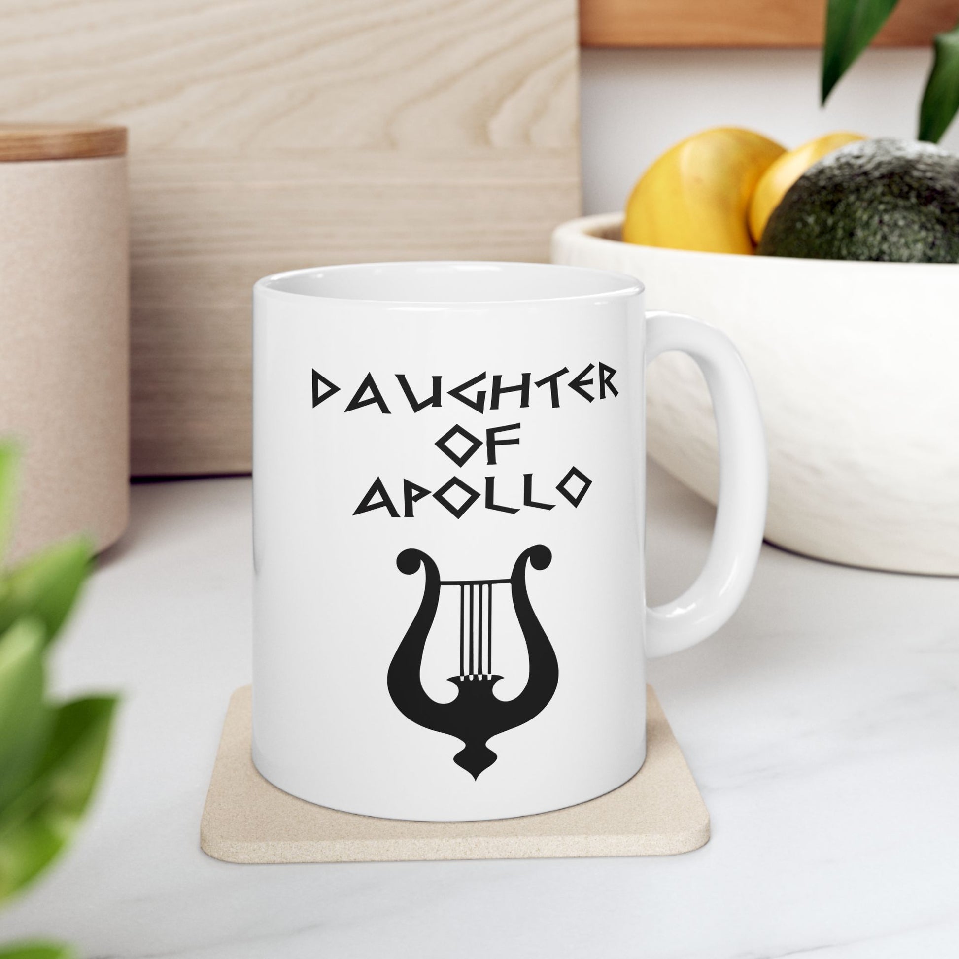 White ceramic mug featuring a minimalist, Greek-inspired graphic in black ink on a white background. The design reads 'DAUGHTER OF APOLLO' in a stylized, angular font, positioned above a clean, line-art graphic of a classical lyre.