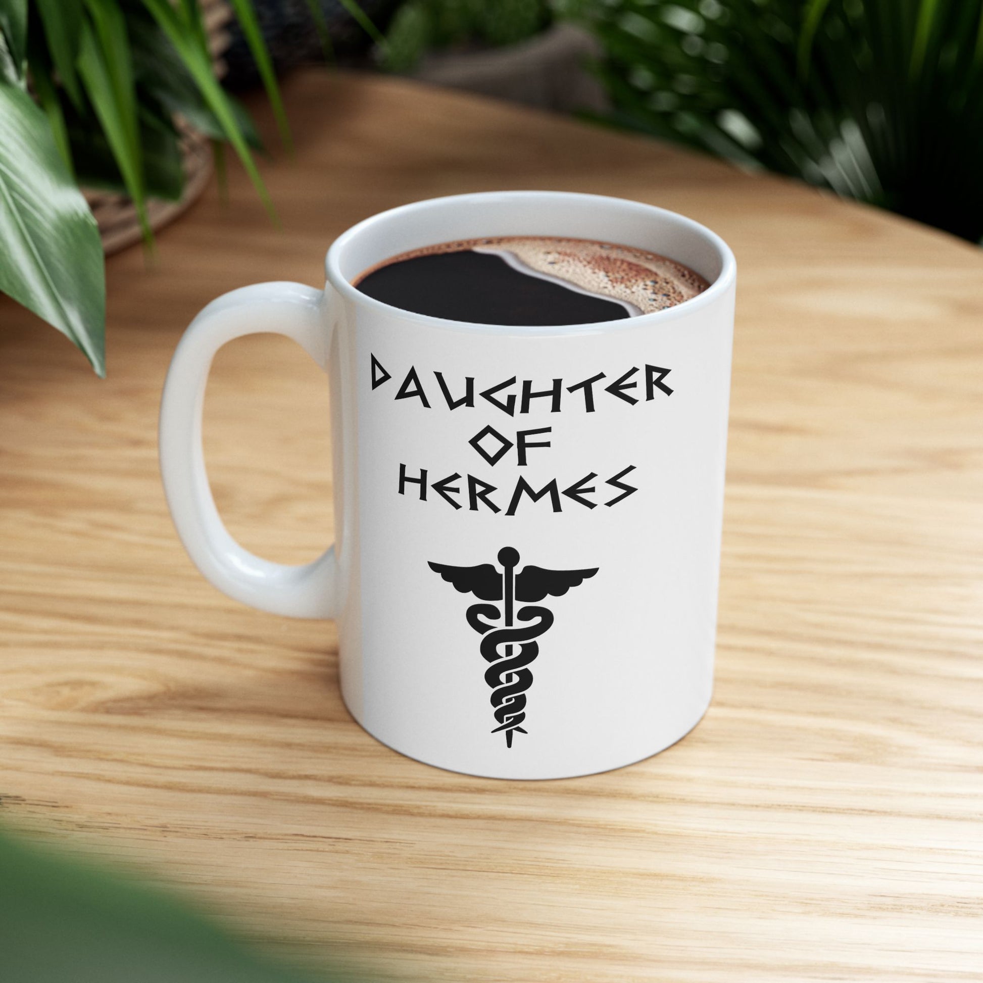 A minimalist black graphic for a white ceramic mug featuring a central caduceus symbol—a winged staff with two snakes entwined around it. Above the symbol, the text "DAUGHTER OF HERMES" is displayed in a sharp, angular font inspired by ancient Greek stone carvings. The entire design is rendered in a clean, high-contrast black outline.