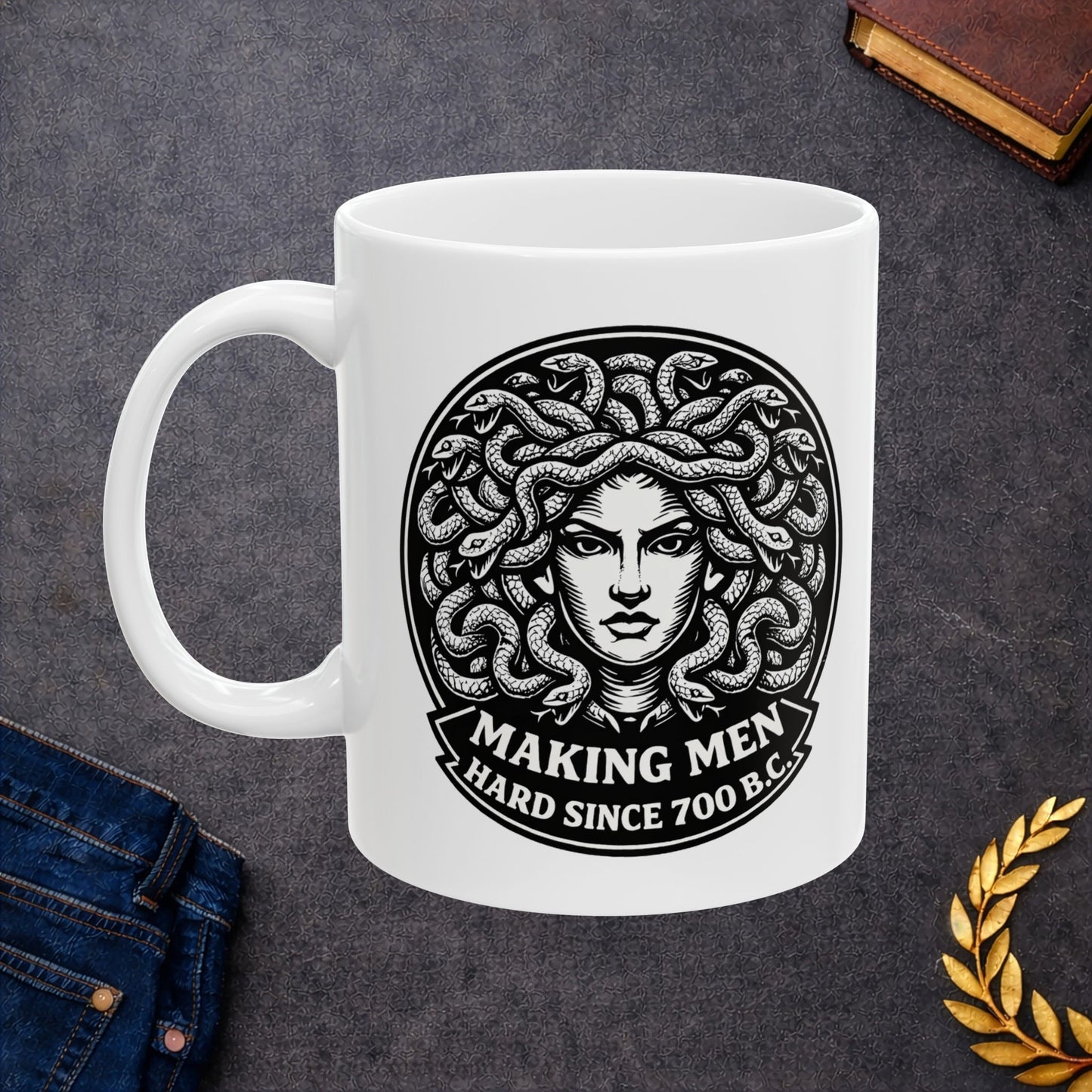 White ceramic mug with a bold black-and-white circular emblem featuring the head of Medusa with a mass of detailed, writhing snakes for hair. Below the illustration, a curved banner contains the humorous double-entendre "MAKING MEN HARD SINCE 700 B.C." written in a heavy, white serif font. The design uses a high-contrast, woodcut-inspired aesthetic with intricate line work against a dark background.