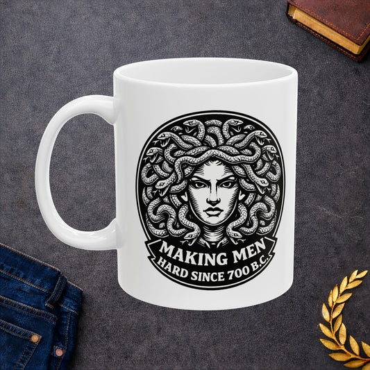 White ceramic mug with a bold black-and-white circular emblem featuring the head of Medusa with a mass of detailed, writhing snakes for hair. Below the illustration, a curved banner contains the humorous double-entendre "MAKING MEN HARD SINCE 700 B.C." written in a heavy, white serif font. The design uses a high-contrast, woodcut-inspired aesthetic with intricate line work against a dark background.