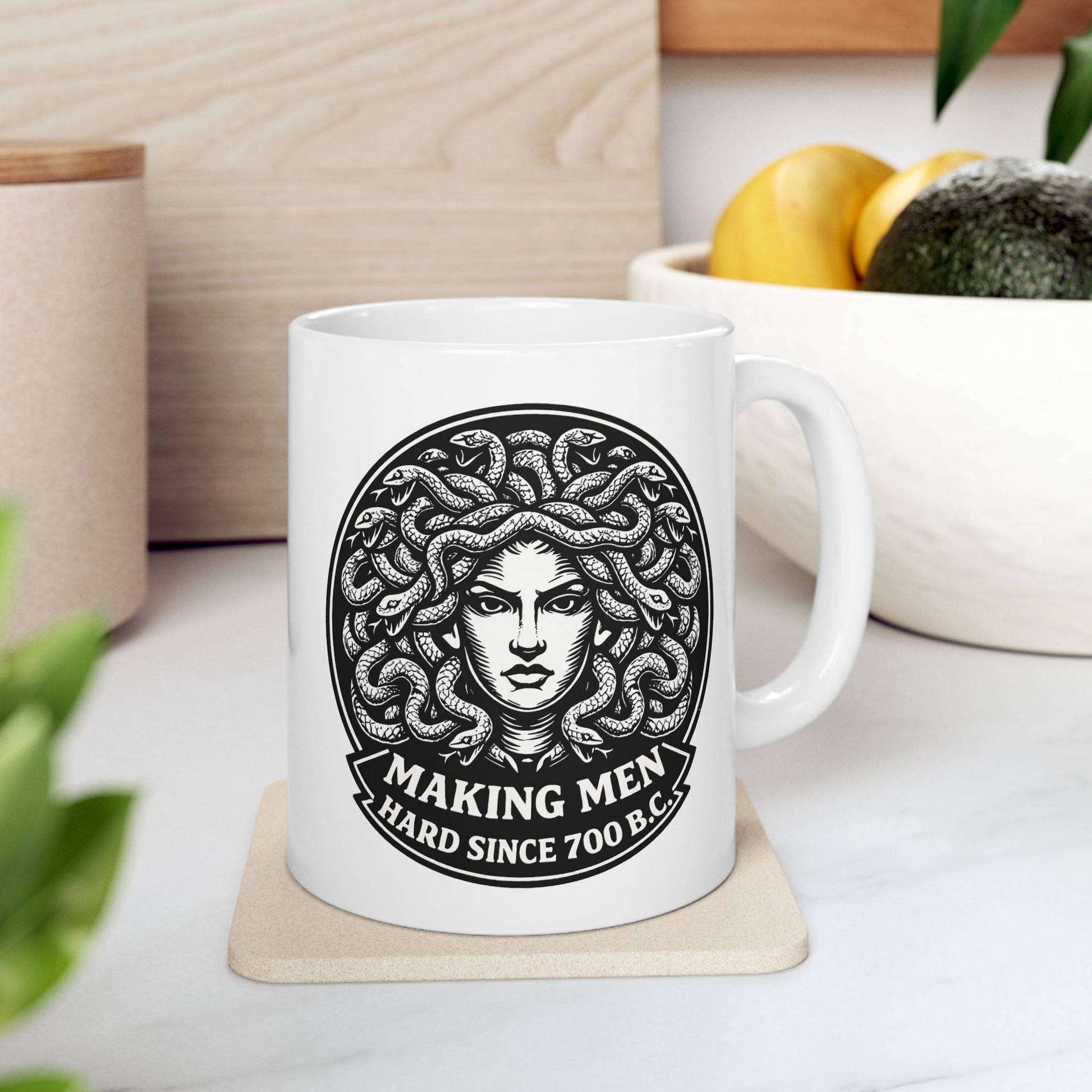 White ceramic mug with a bold black-and-white circular emblem featuring the head of Medusa with a mass of detailed, writhing snakes for hair. Below the illustration, a curved banner contains the humorous double-entendre "MAKING MEN HARD SINCE 700 B.C." written in a heavy, white serif font. The design uses a high-contrast, woodcut-inspired aesthetic with intricate line work against a dark background.
