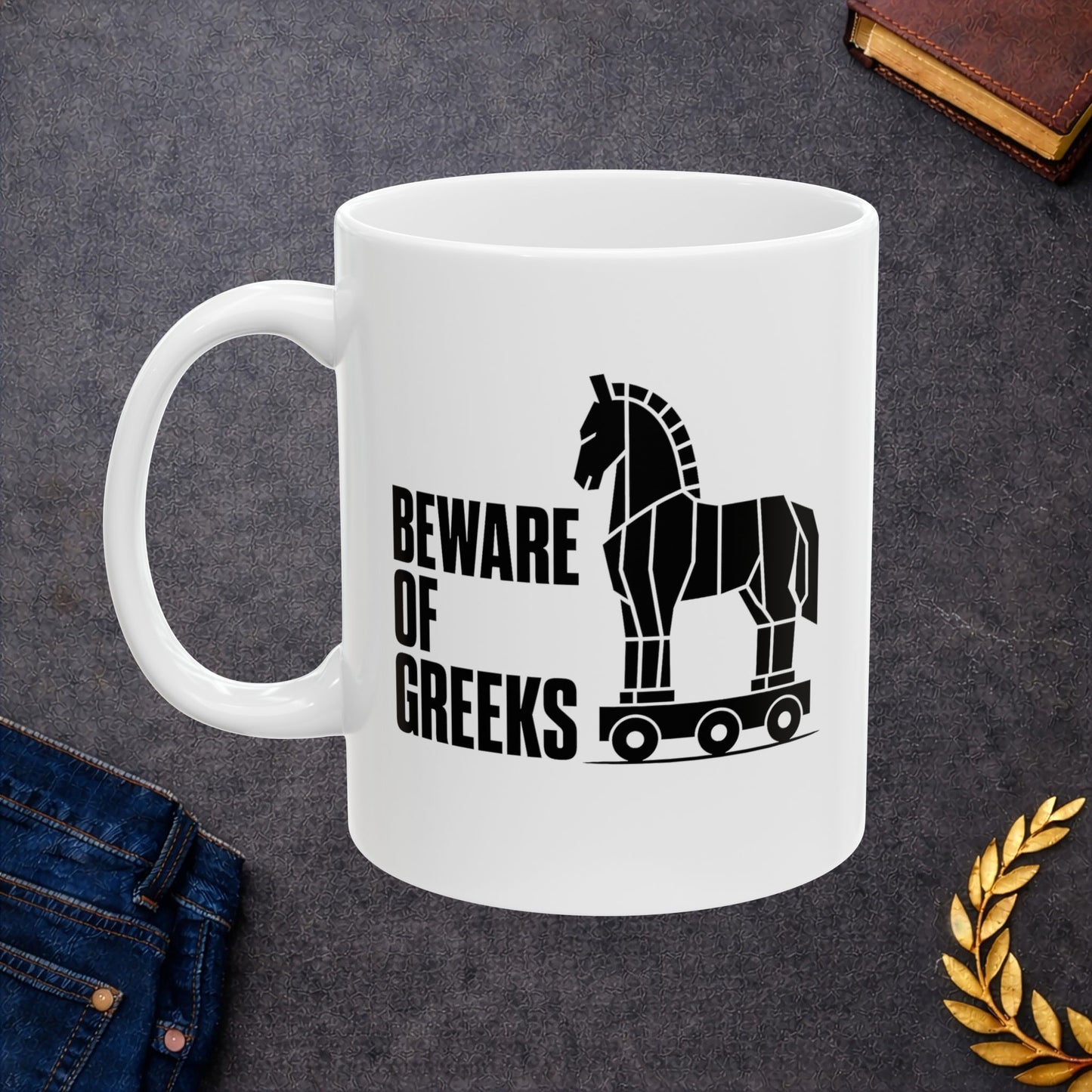 White ceramic mug with a minimalist black-and-white graphic featuring a stylized, geometric silhouette of the Trojan Horse on a wheeled platform. To the left of the horse, the text "BEWARE OF GREEKS" is written vertically in a bold, stacked, black sans-serif font. The design uses clean lines and high contrast on a plain background, offering a modern, minimalist take on the classic Greek mythological tale.