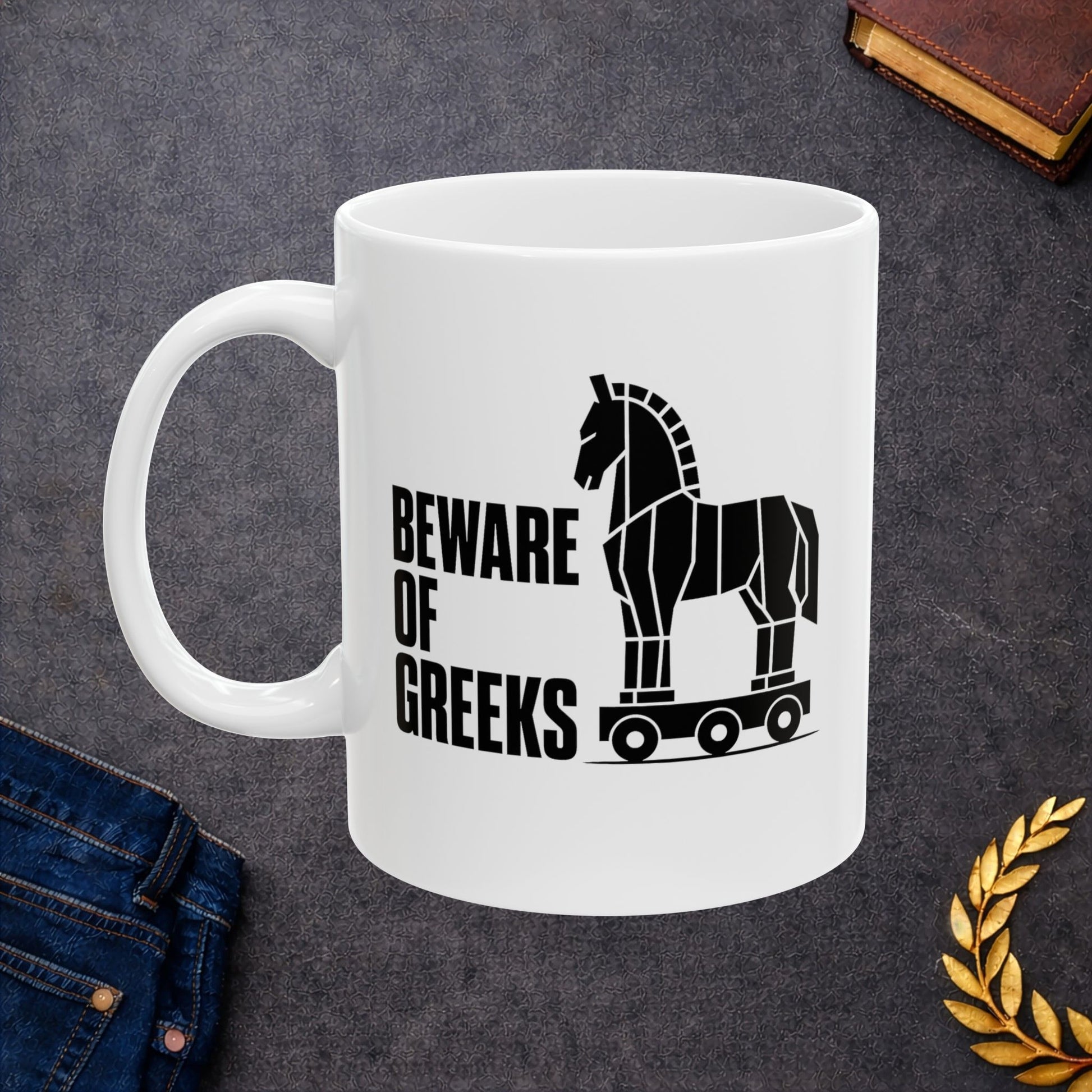White ceramic mug with a minimalist black-and-white graphic featuring a stylized, geometric silhouette of the Trojan Horse on a wheeled platform. To the left of the horse, the text "BEWARE OF GREEKS" is written vertically in a bold, stacked, black sans-serif font. The design uses clean lines and high contrast on a plain background, offering a modern, minimalist take on the classic Greek mythological tale.