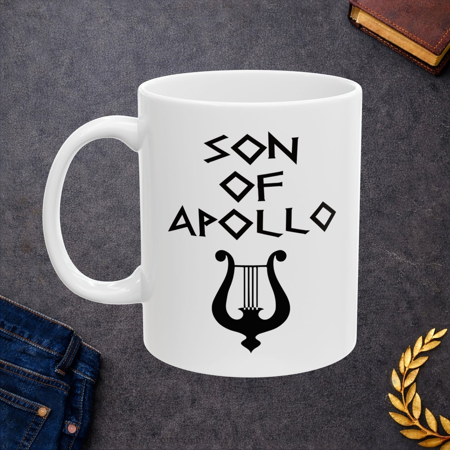 White ceramic mug featuring a minimalist, Greek-inspired graphic in black ink on a white background. The design reads 'SON OF APOLLO' in a stylized, angular font, positioned above a clean, line-art graphic of a classical lyre.