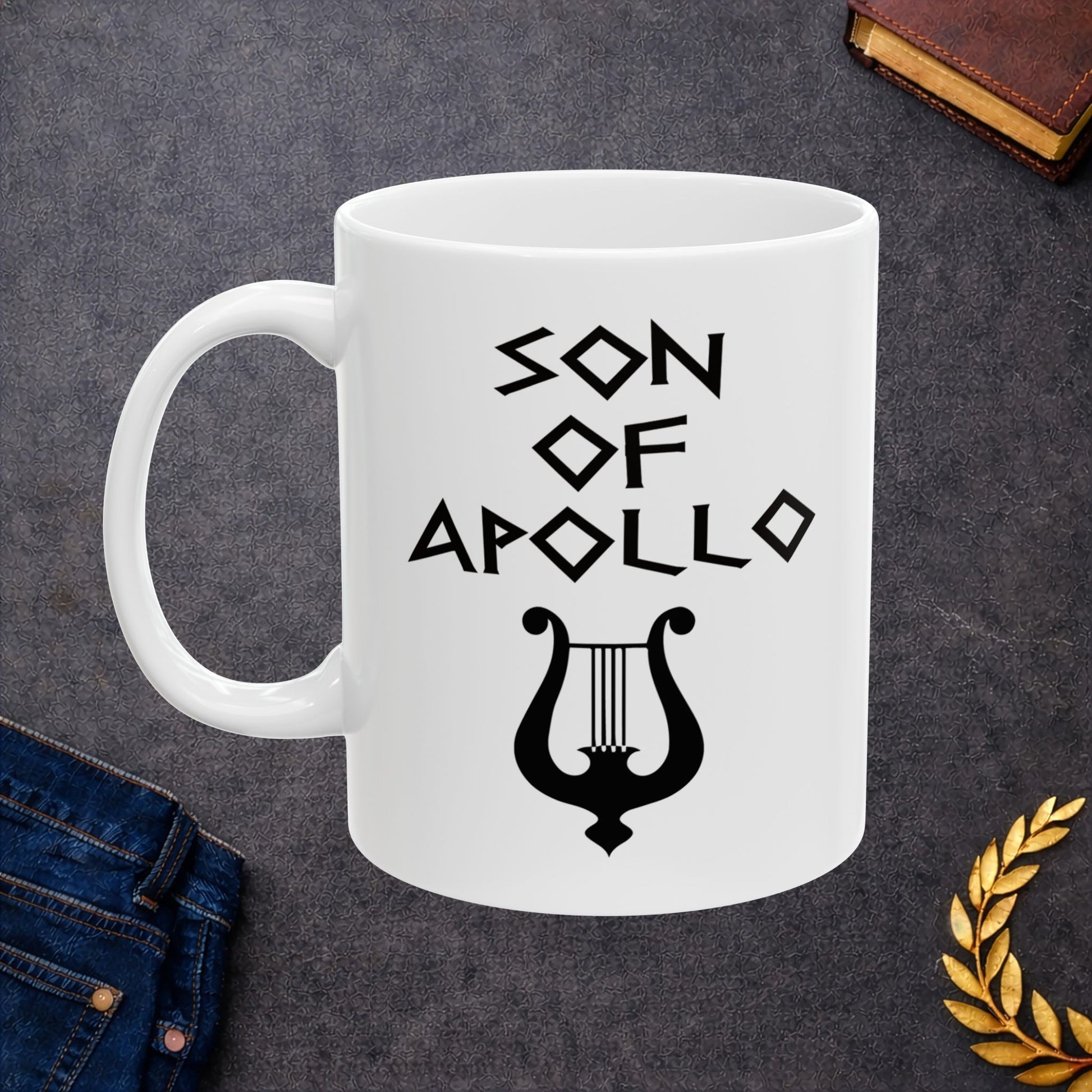 White ceramic mug featuring a minimalist, Greek-inspired graphic in black ink on a white background. The design reads 'SON OF APOLLO' in a stylized, angular font, positioned above a clean, line-art graphic of a classical lyre.